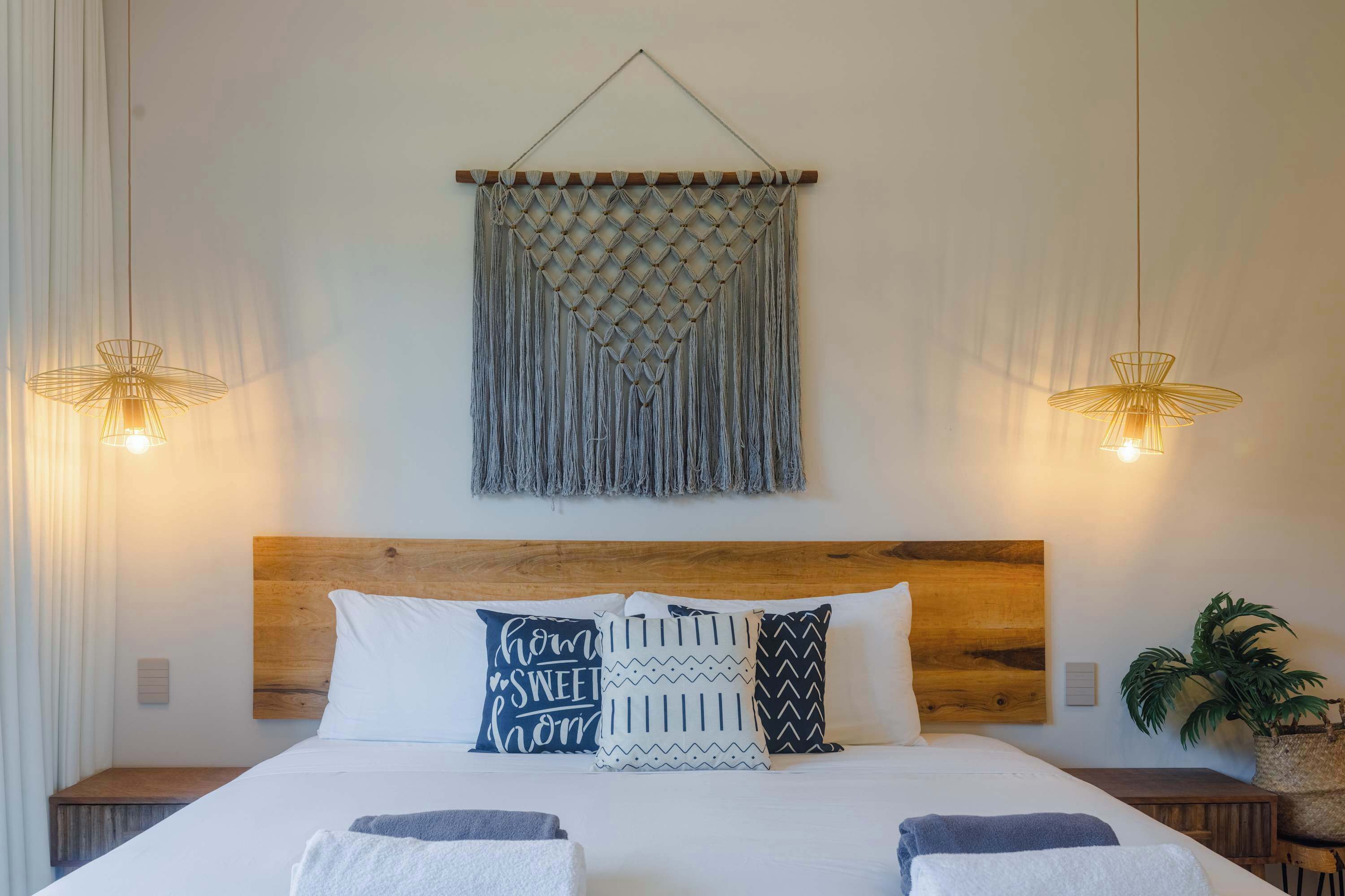 A neatly made bed with a wooden headboard, a macrame wall hanging, and two wall-mounted lamps.