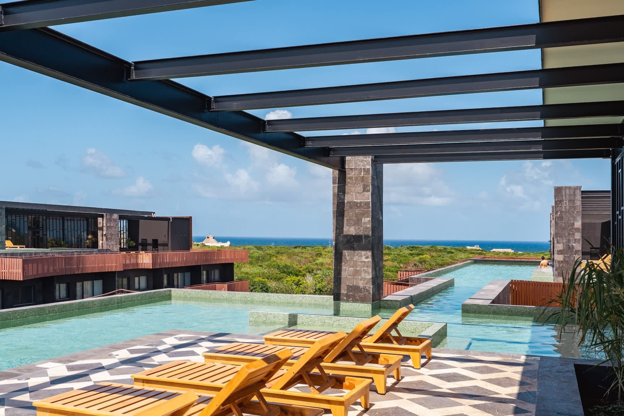 Una piscina infinita en la azotea con tumbonas de madera, vistas despejadas al océano y una estructura de edificio moderna.