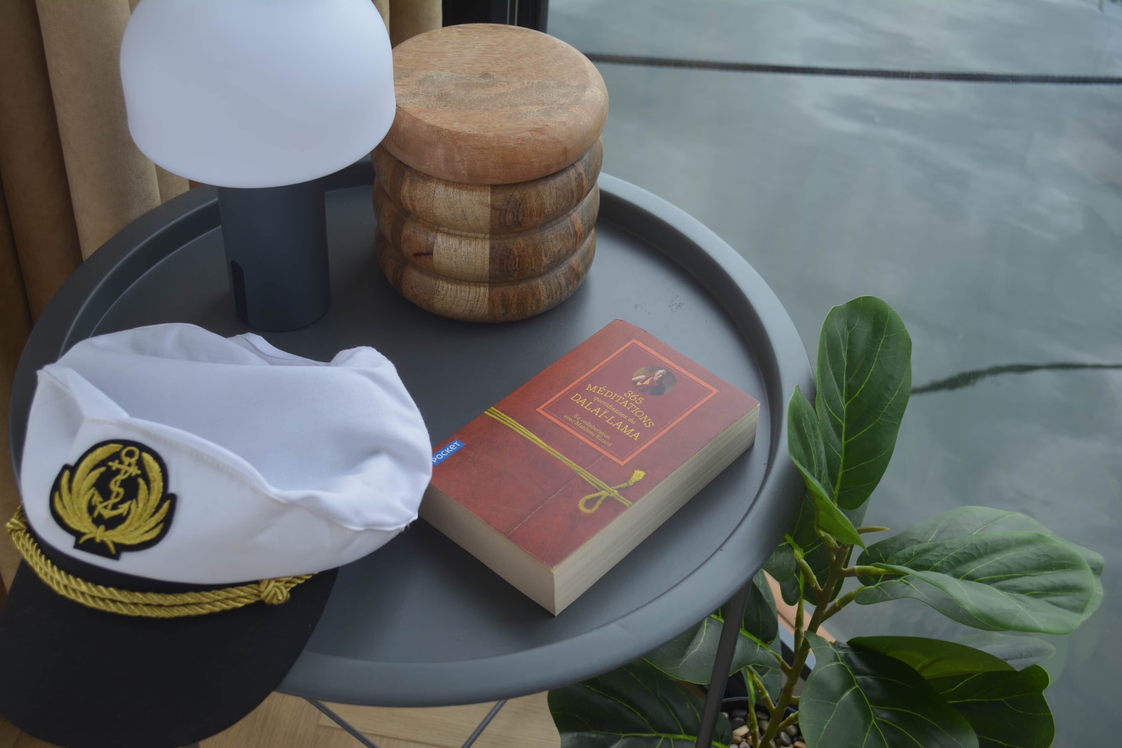 Um boné branco com emblema dourado, um livro intitulado '365 MEDITAÇÕES DALAI-LAMA' e um banquinho de madeira sobre uma mesa redonda.