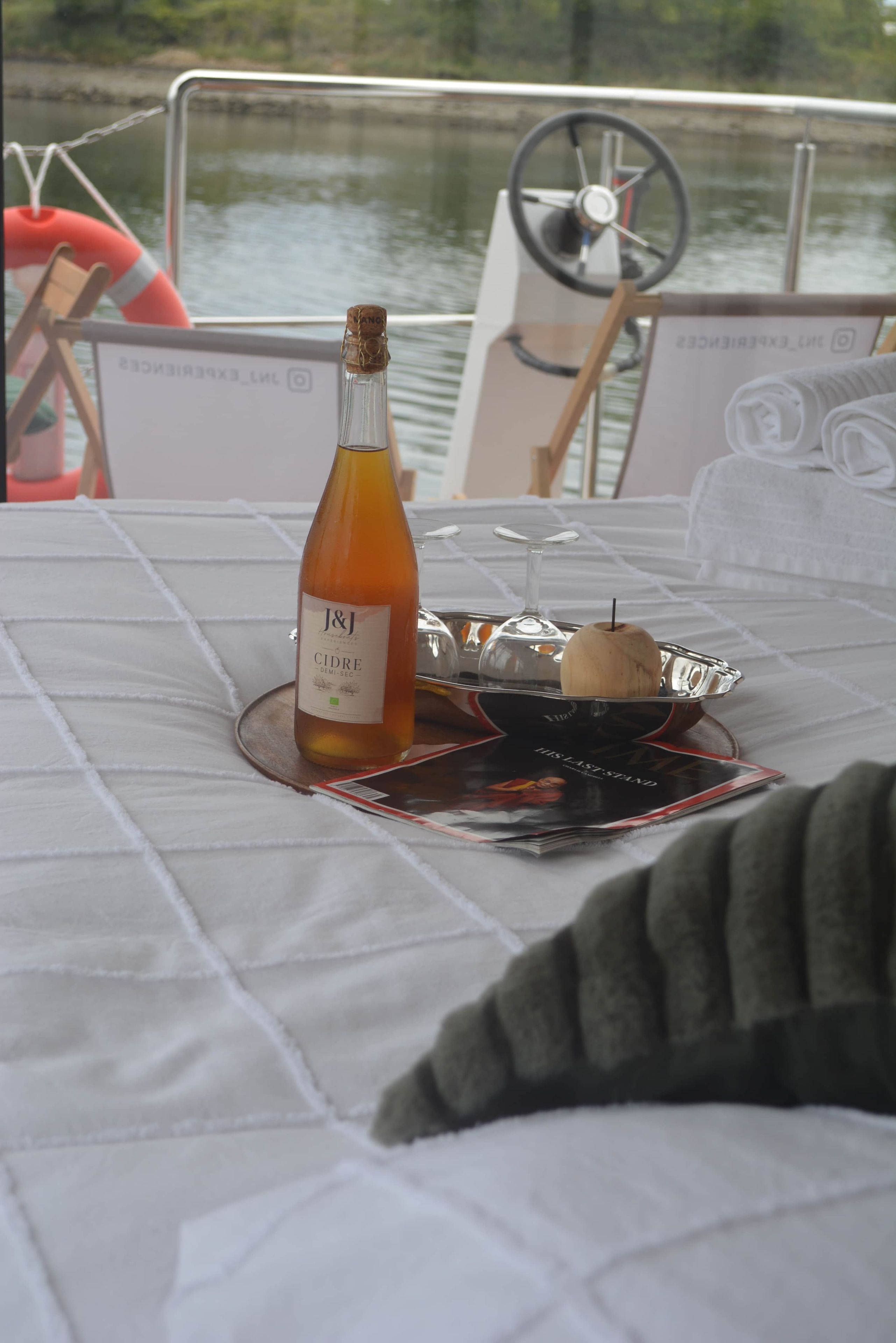 Ein Tisch mit einer Flasche J&J Wein, einem Glas und einer Schale mit einem Holzgegenstand, vor dem Hintergrund von Wasser und dem Steuerrad eines Bootes.
