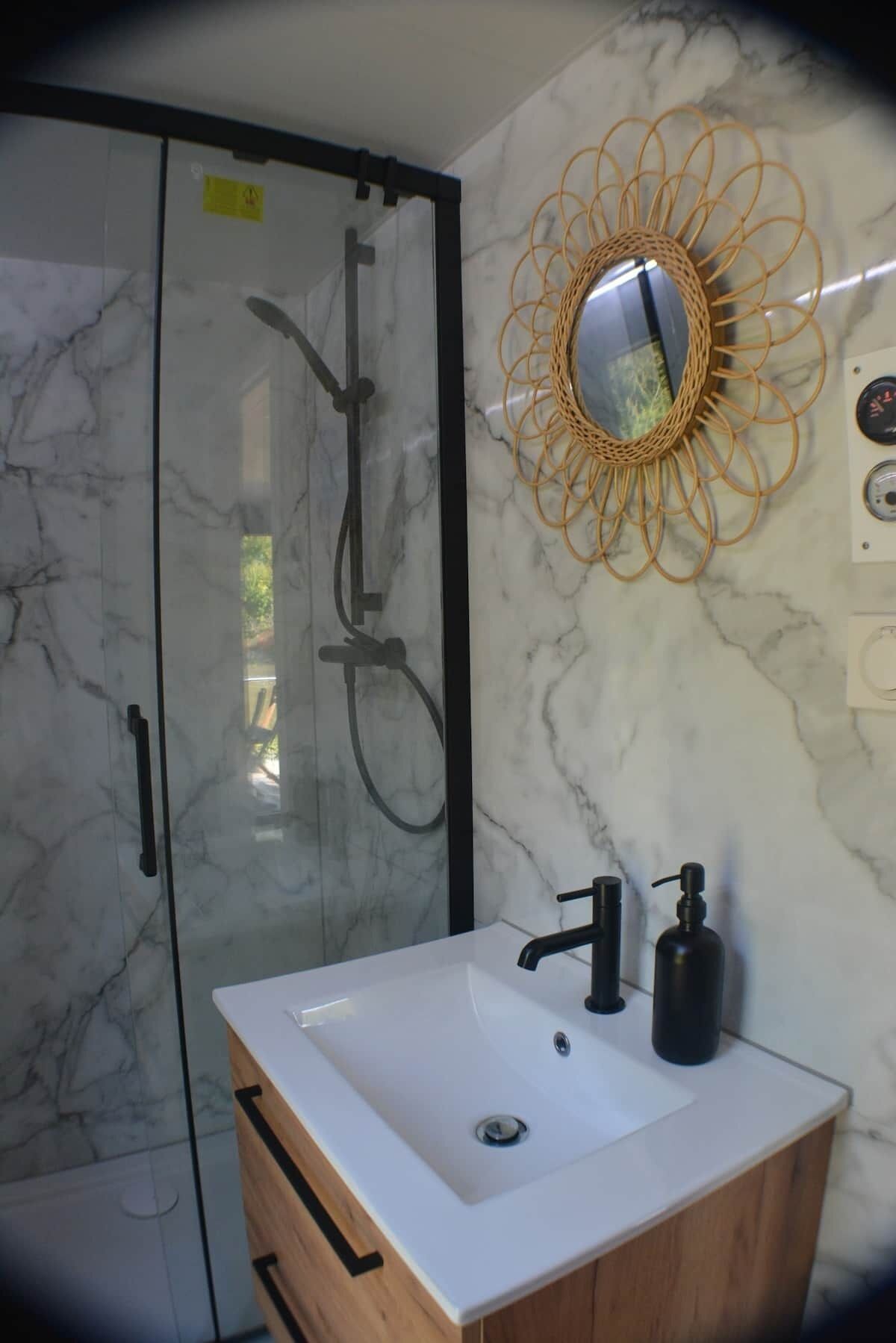 Un baño con cabina de ducha de vidrio, un lavabo blanco con grifo negro y un espejo decorativo en la pared.