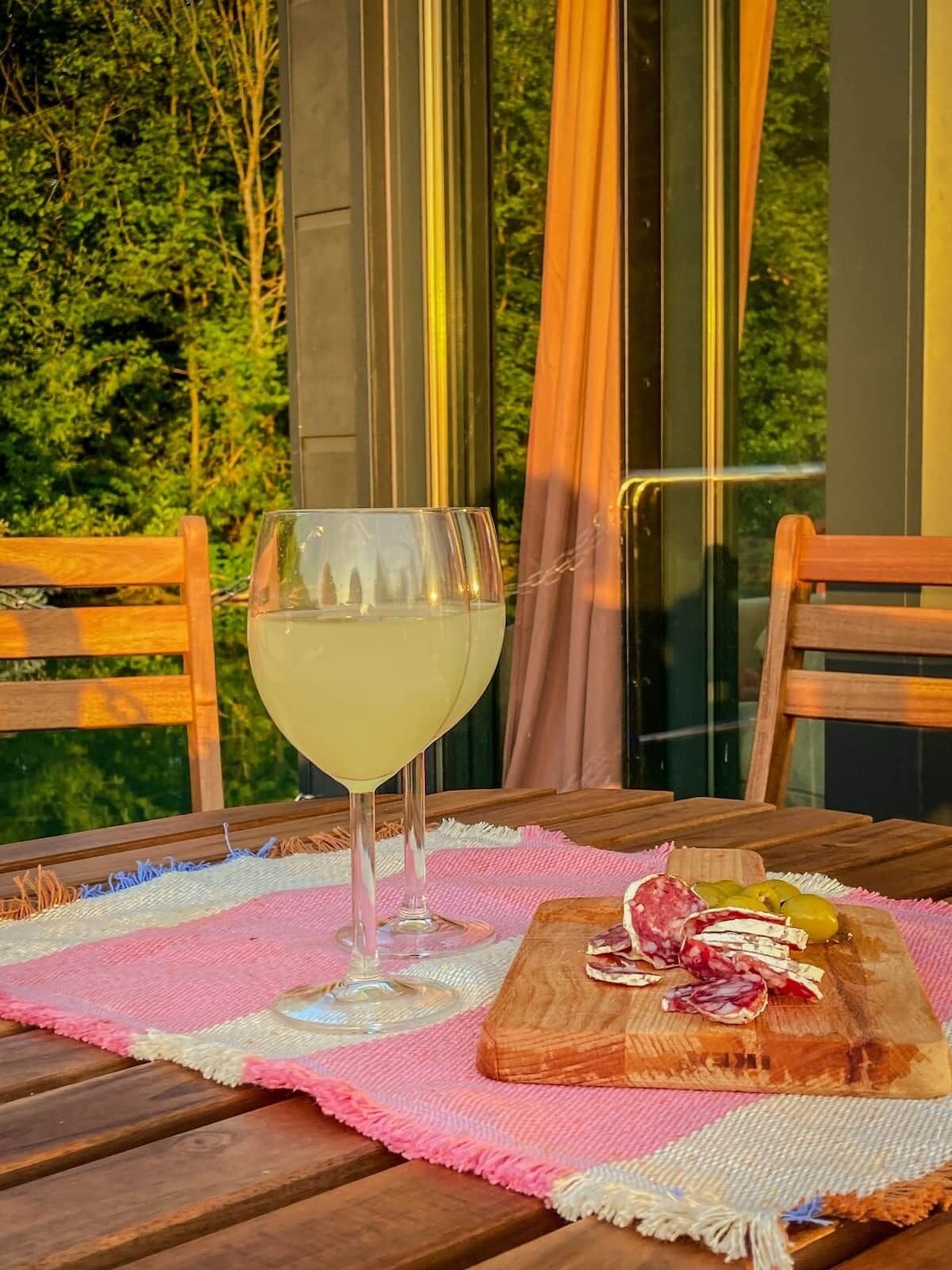 Una mesa con una copa de vino blanco, una tabla de madera con varios quesos y aceitunas, y un mantel tejido en rosa y blanco.
