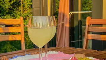 Una mesa con una copa de vino blanco, una tabla de madera con varios quesos y aceitunas, y un mantel tejido en rosa y blanco.
