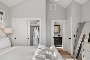 Quarto com cama, mesa de cabeceira, abajur, guarda-roupa e banheiro visível por uma porta aberta.