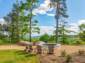 Uma fogueira rodeada por cadeiras Adirondack numa área arborizada, com vista para montanhas.