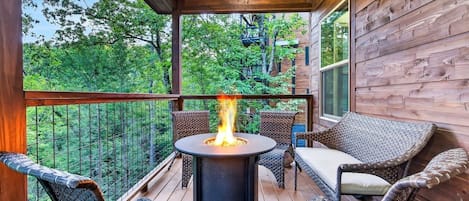 A wooden deck with a round table and chairs, a fire pit, and a view of trees.
