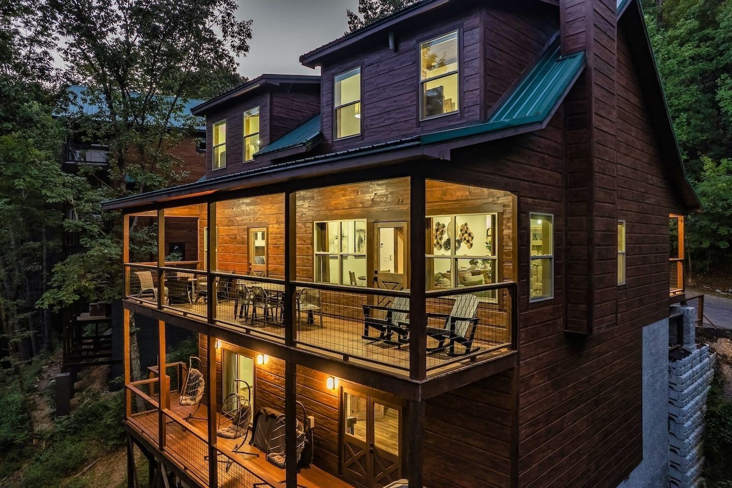 A two-story wooden cabin with a balcony, large windows, and a view of the surrounding forest.