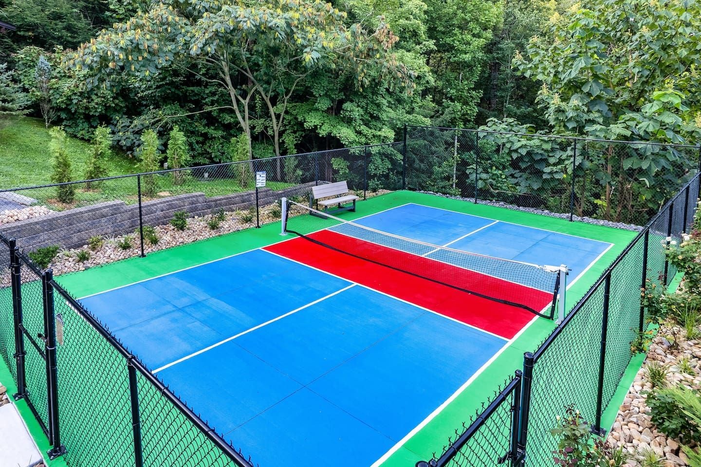 Un terrain de tennis en plein air, entouré d’une clôture en treillis métallique, avec un banc et des arbres en arrière-plan.