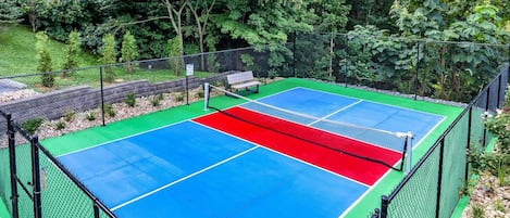 Cancha de tenis al aire libre rodeada de una cerca de malla metálica, con un banco y árboles al fondo.