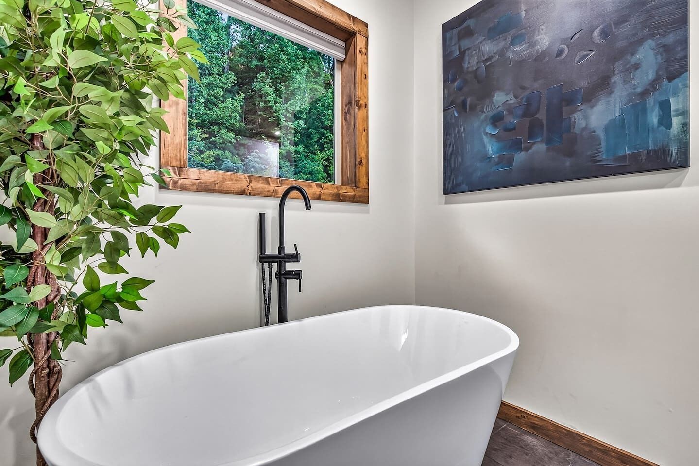 A bathroom with a freestanding bathtub, a window with a view of greenery, and a painting on the wall.