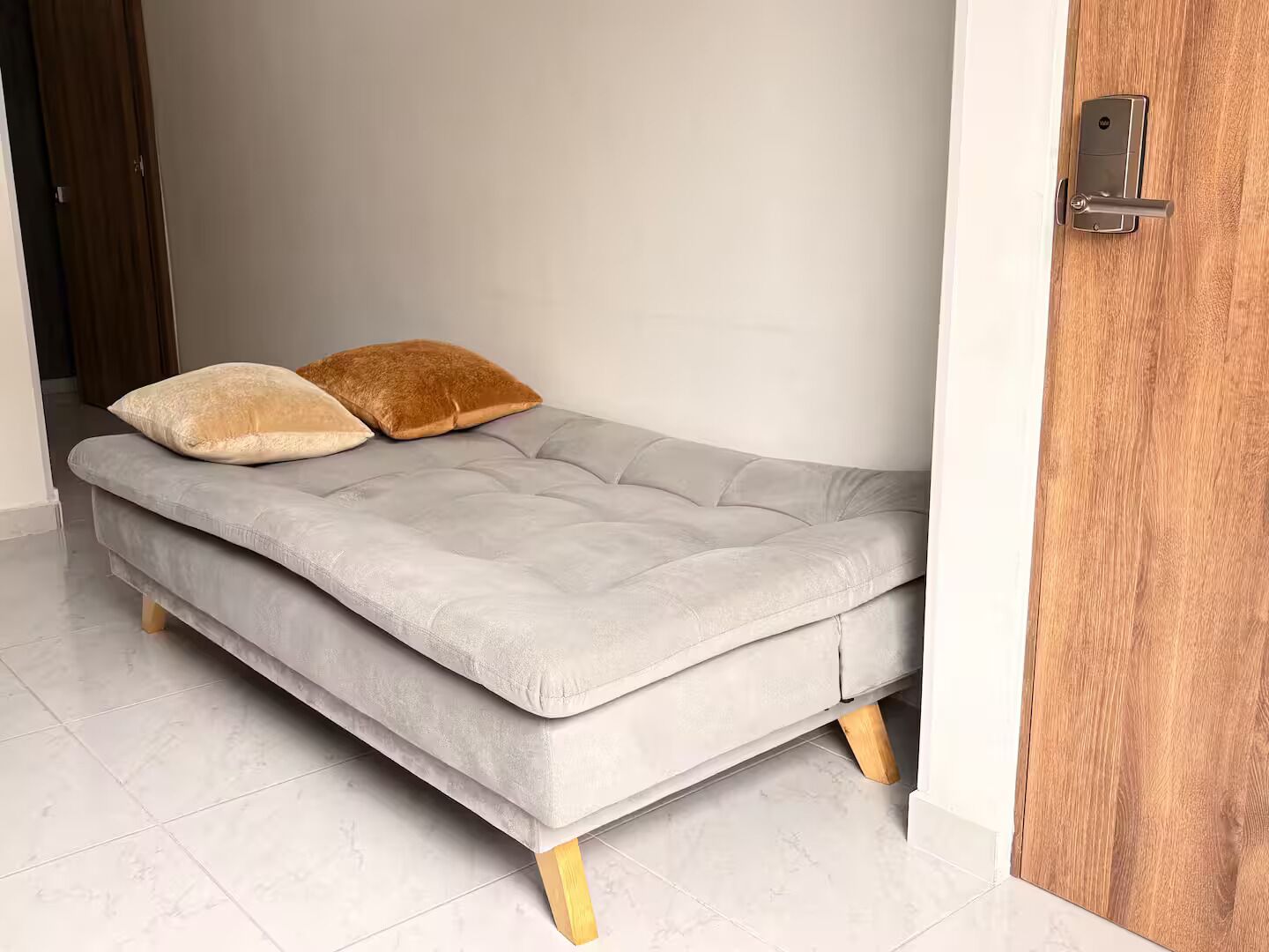 A single bed with a grey upholstered headboard and wooden legs, accompanied by two pillows, in a room with a wooden door and white walls.