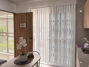 Ein modernes Zimmer mit einer Holztür mit der Aufschrift '202', einem weißen Vorhang mit vertikalen Linien, einer Orchidee im Topf auf einem Tisch sowie einer Küchenzeile mit Spüle und einem violetten Handtuch.