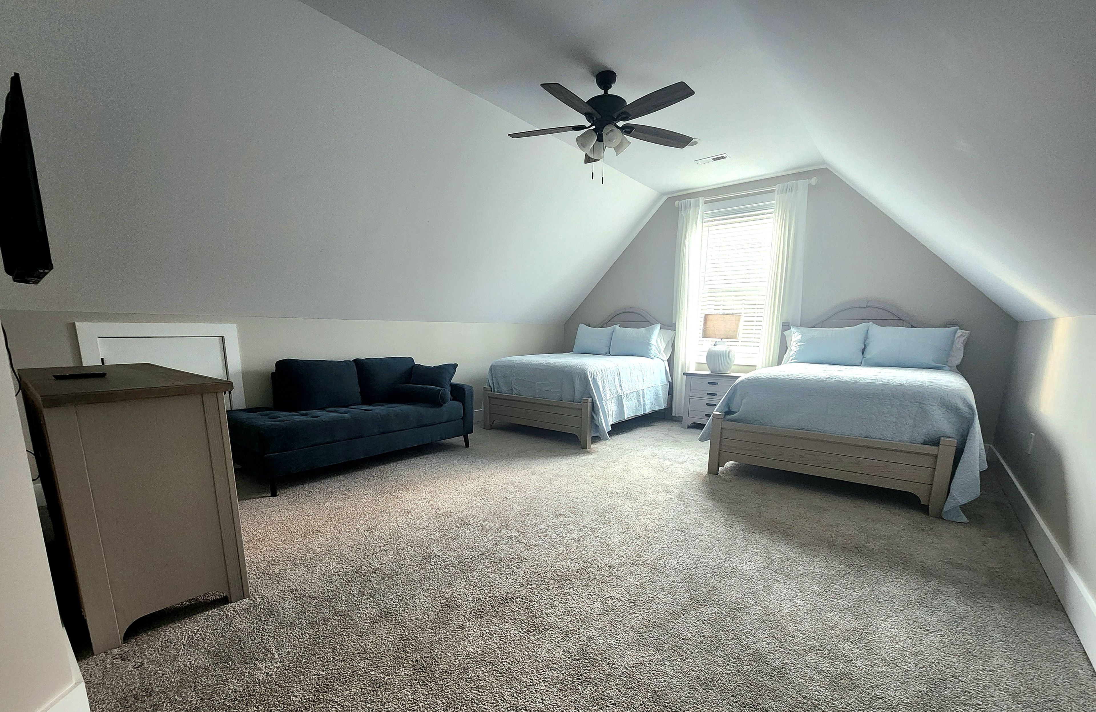A bedroom with two beds, a ceiling fan, a window with blinds, and a dark-colored sofa.