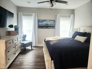 Ein Schlafzimmer mit einem Bett, einem Schminktisch, einem Sessel, einem Deckenventilator und einem Fenster mit Vorhängen.