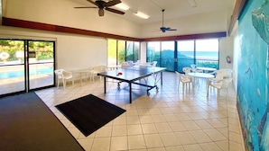 Um quarto amplo com mesa de pingue-pongue, ventiladores de teto, grandes janelas e vista para o oceano.