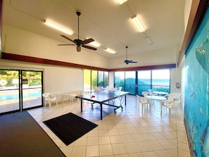 A spacious room with a ping pong table, ceiling fans, large windows, and a view of the ocean.