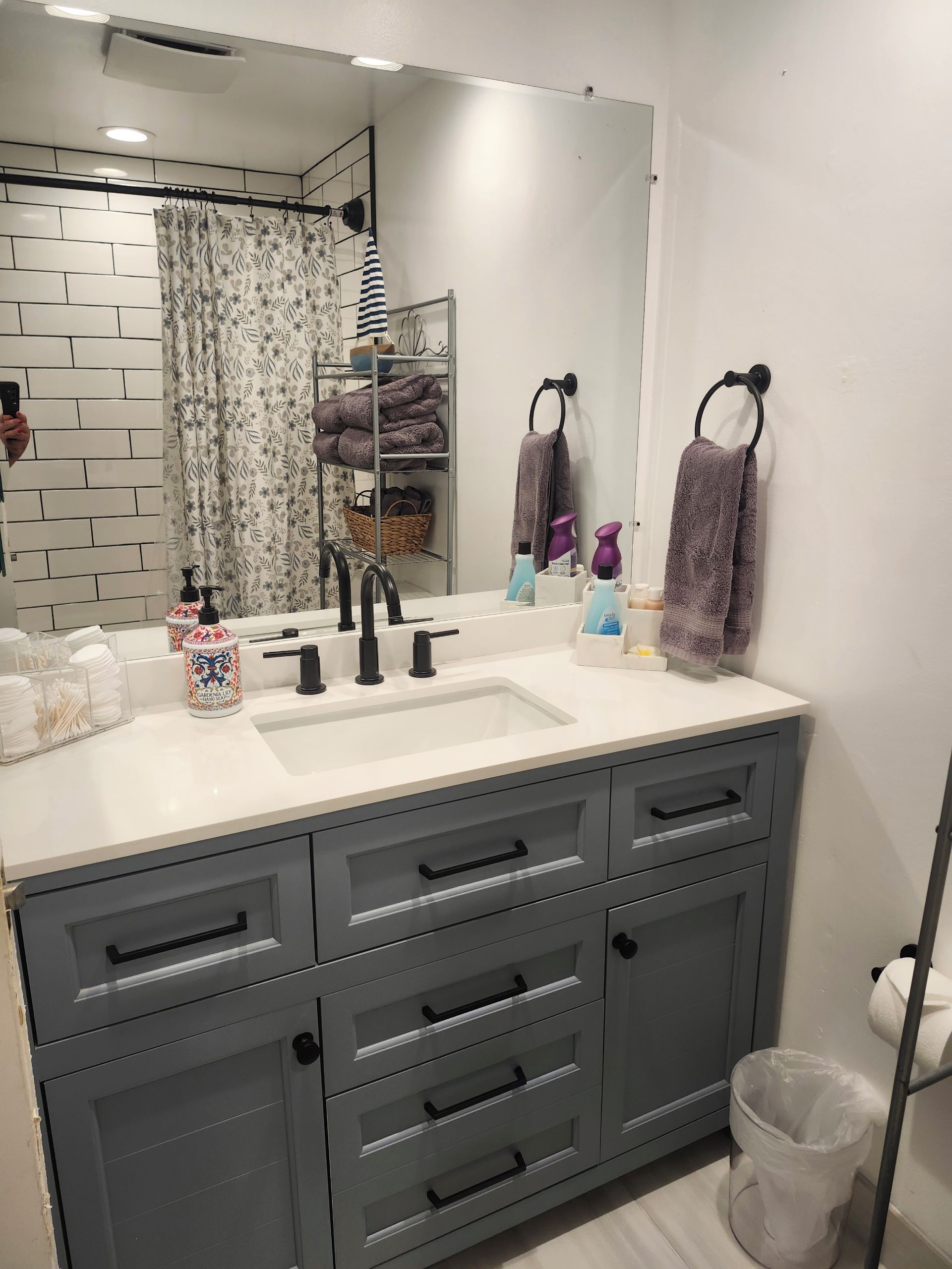 A bathroom with a white sink, grey vanity, and a mirror. There are towels, toiletries, and a trash can.