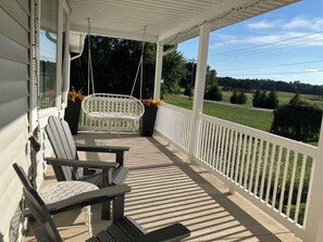 A porch with a swing chair, a table, and chairs, overlooking a green landscape.