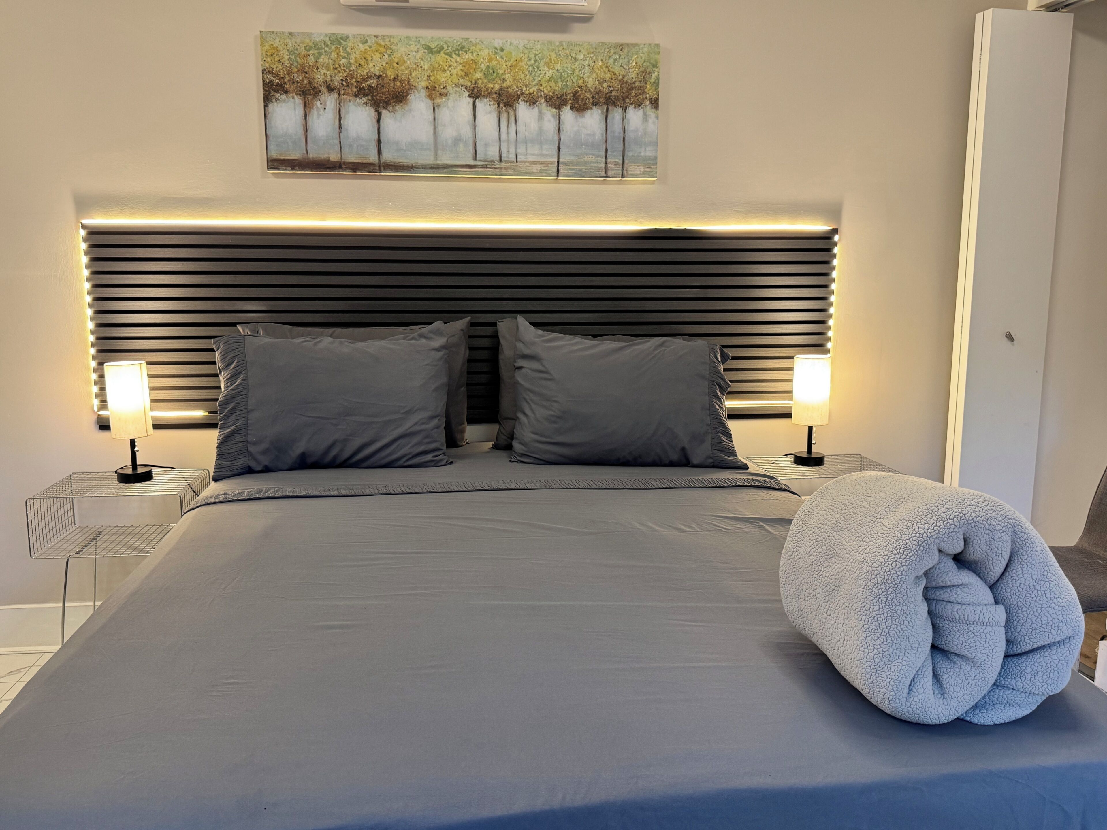 Ein modernes Schlafzimmer mit einem großen Bett, zwei Nachttischlampen, einem Kopfteil mit horizontalen Lamellen und einem Bild über dem Bett.