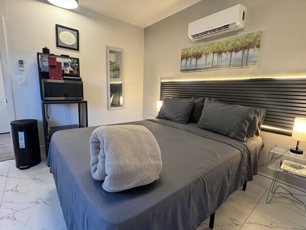 Une chambre d’hôtel comprenant un lit, une table de chevet avec une lampe, une poubelle, un climatiseur fixé au mur et un tableau encadré.