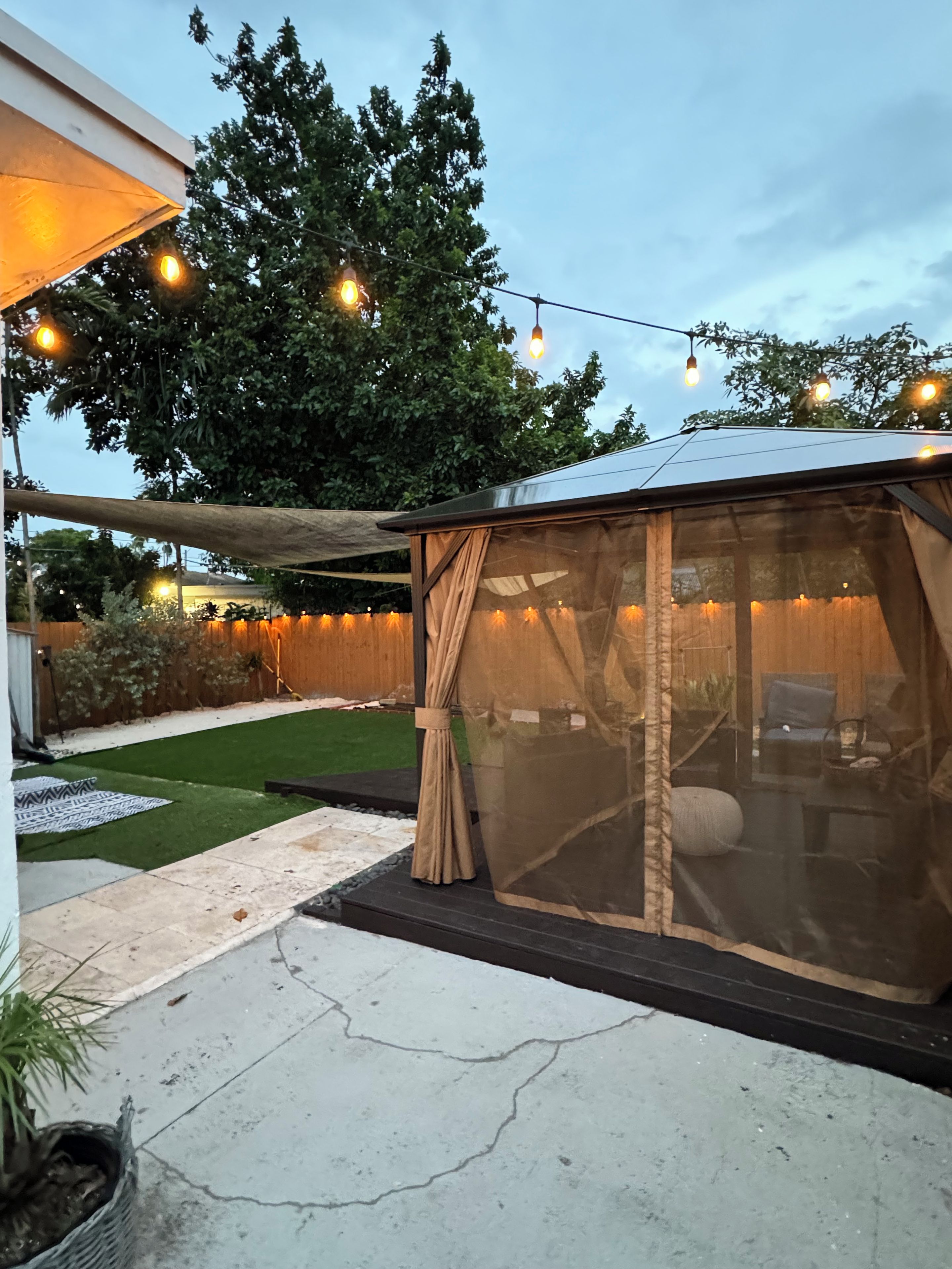 A covered outdoor seating area with string lights, a lawn, and a wooden fence.