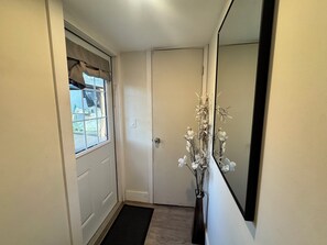 Ein Flur mit einer Tür, einem fensterbehangenen Fenster, einer Fußmatte und einem Spiegel, der eine Vase mit Blumen widerspiegelt.