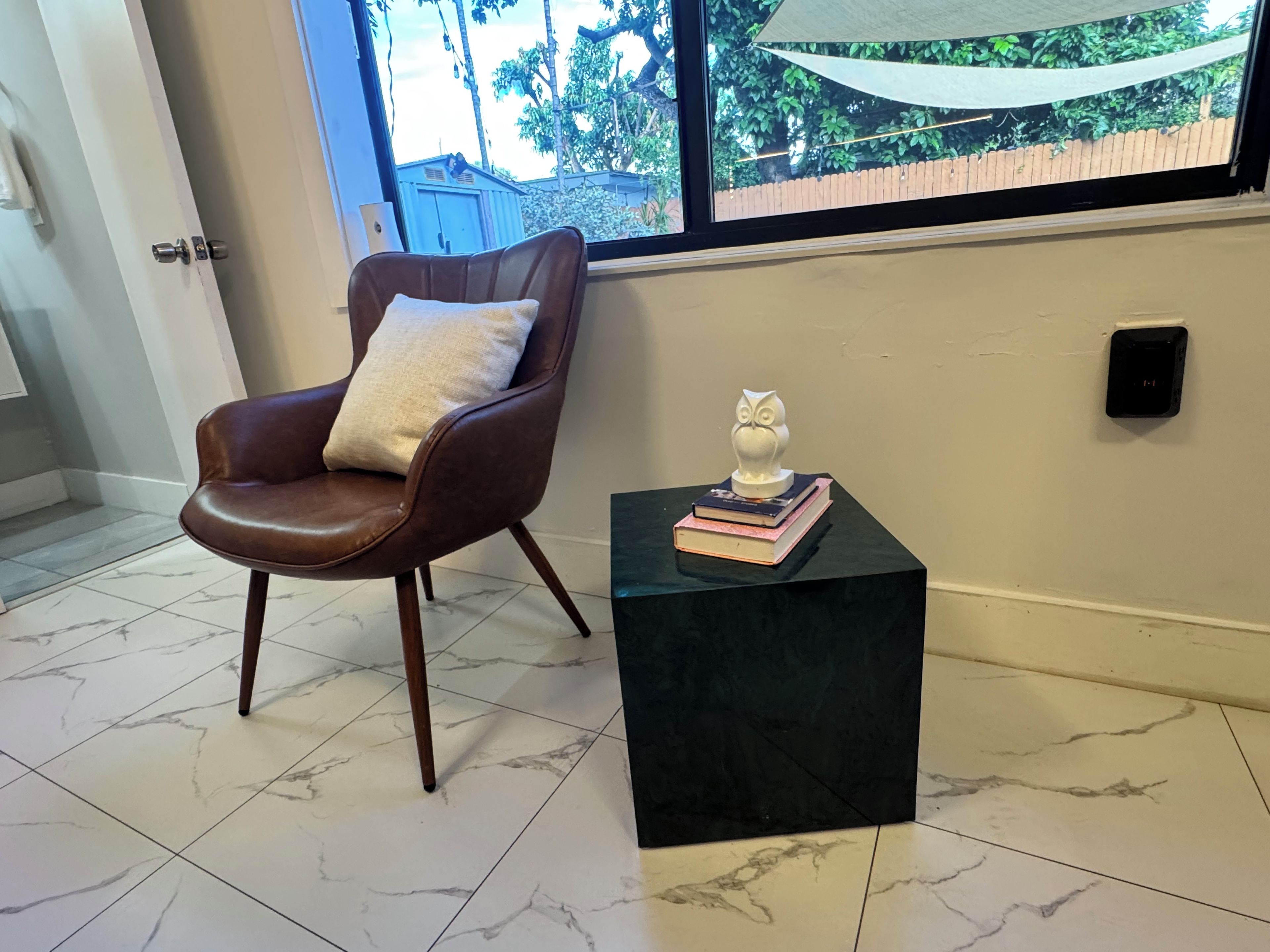 A room with a brown leather chair, a black side table with books and a white owl sculpture, and a window with a view of greenery.