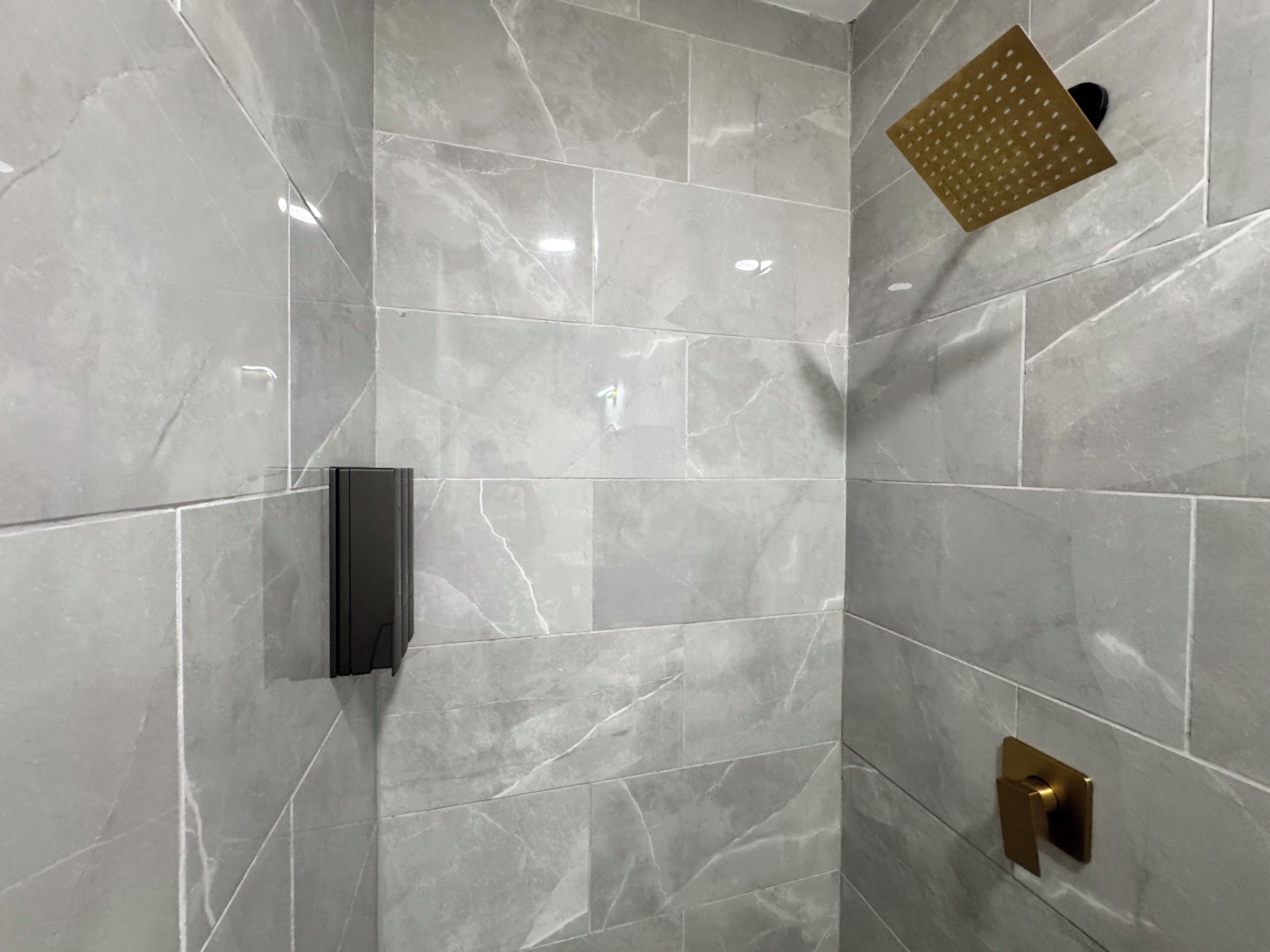 A bathroom with marble tiles, a black towel dispenser, a gold-colored shower control, and a handheld showerhead.