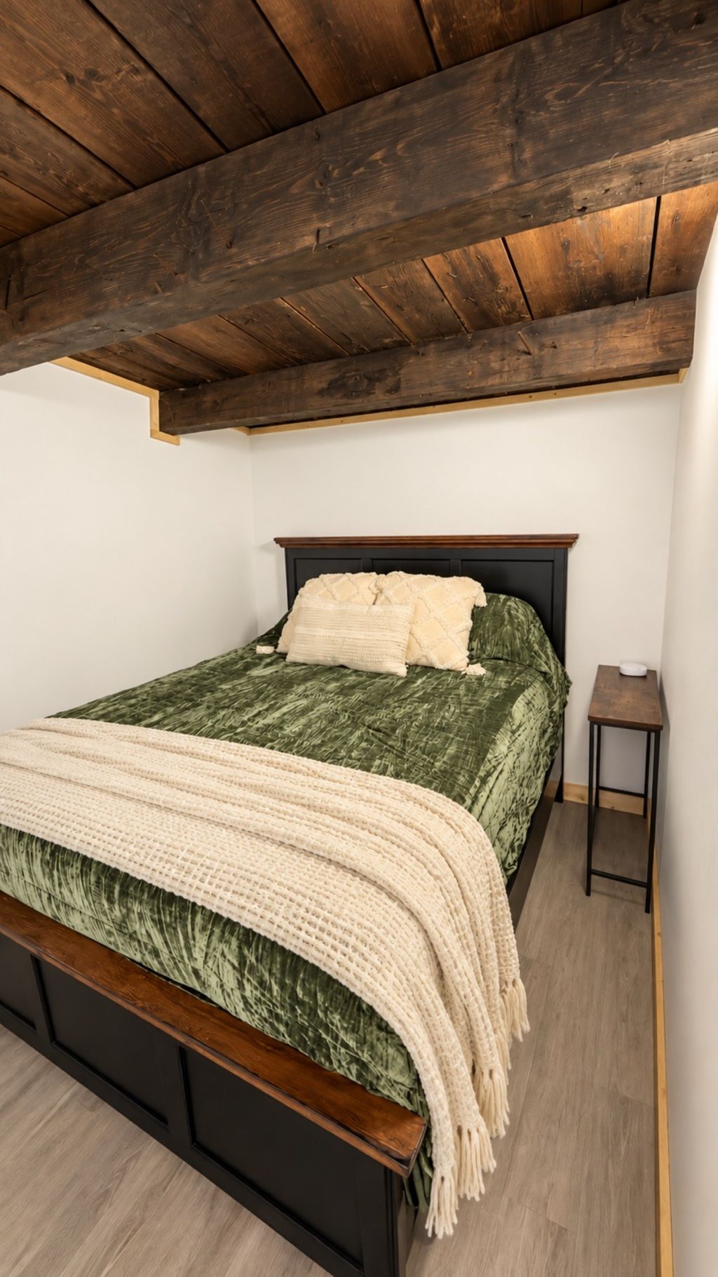 Un dormitorio con techo de madera, una cama con colcha verde y almohadas blancas, una mesita de noche de madera y una pared blanca.
