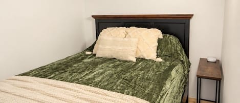 Ein Schlafzimmer mit Holzboden, einem Bett mit grüner Bettdecke und weißen Kissen, einem Nachttisch aus Holz und einer weißen Wand.