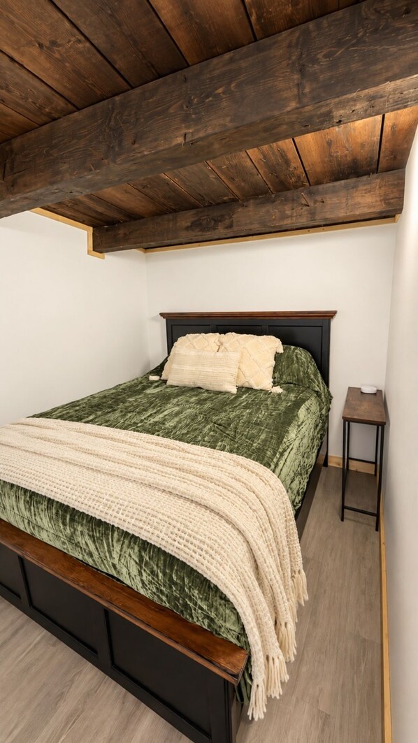 Une chambre avec un plafond en bois, un lit recouvert d’une couverture verte et agrémenté d’oreillers blancs, une table de chevet en bois et un mur blanc.