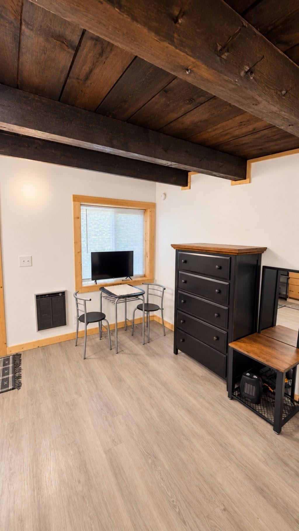 A room with a wooden ceiling, a window with blinds, a television, a desk with two chairs, a black dresser, and a small table with a speaker.