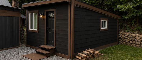 Ein kleines, dunkelholziges Gebäude mit Tür und Fenstern, eine Schotterauffahrt und ein Holzstapel.