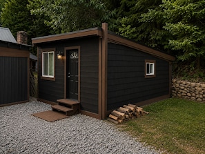 A small, dark-colored wooden structure with a door and windows, a gravel driveway, and a stack of firewood.