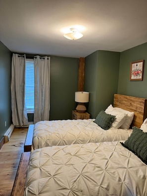 A bedroom with two beds, a nightstand with a lamp, a wooden headboard, and a window with curtains. - Charming First Floor Farmhouse (Clarendon)
