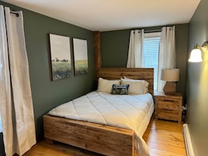 A bedroom with a wooden bed, a nightstand with a lamp, and two wall-mounted paintings. - Charming First Floor Farmhouse (Clarendon)
