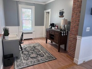 A room with a window, a radiator, a desk with a chair, a rug, and a brick wall.