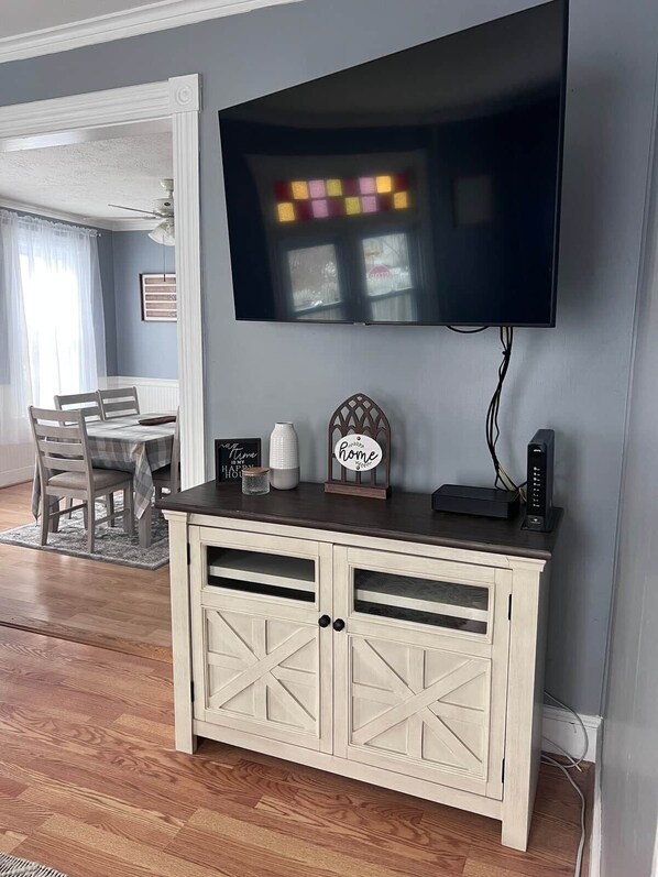 A living room with a flat-screen TV mounted on the wall, a wooden cabinet with drawers, a dining table with chairs, and a rug on the floor.