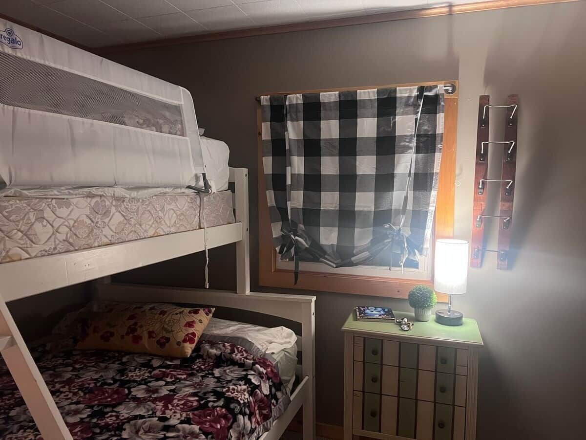 A dormitory room with bunk beds, a window with checkered curtains, a desk with a lamp, and a small cabinet.