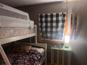 A dormitory room with bunk beds, a window with checkered curtains, a desk with a lamp, and a small cabinet. - Reflections on the Lake (Hampton)