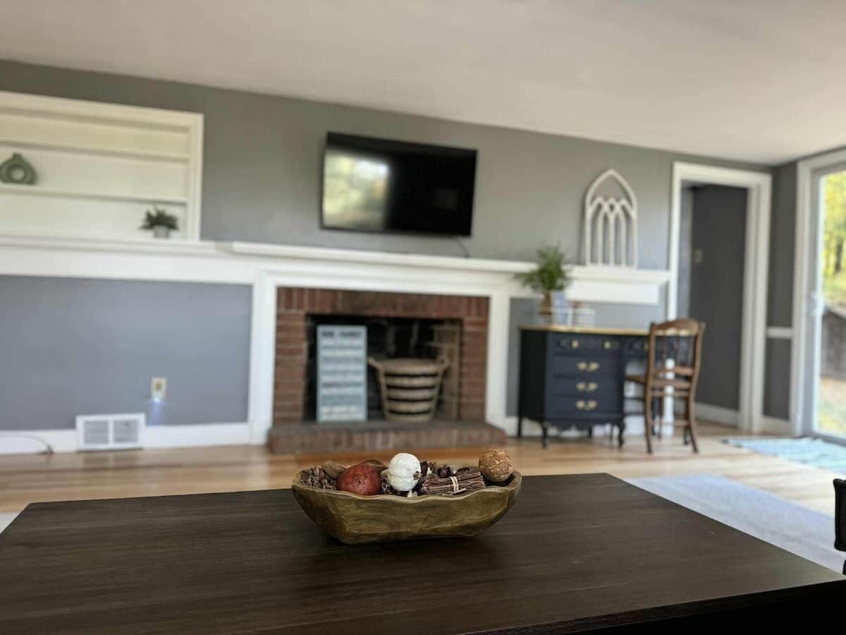 A living room with a fireplace, a television mounted above it, and a dining table with a bowl of snacks in the foreground.
