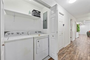 A laundry room with a front-loading washer and dryer, a shelf with laundry baskets, and a door leading to another room. - Palmetto Safari Retreat (Horry County)