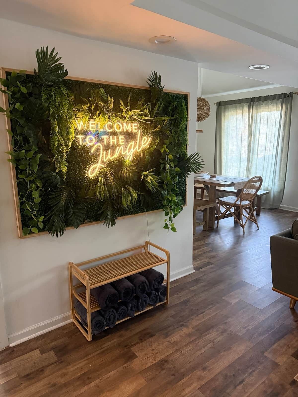 A living room with a wall-mounted sign that reads 'WELCOME TO THE Jungle,' a dining area with a wooden table and chairs, and a wooden bench with rolled-up mats.