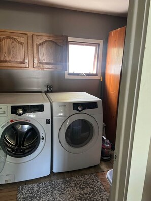 A laundry room with two white Samsung washing machines, wooden cabinets above, a window, and a patterned rug. - Quaint City Cottage just minutes from Killington (Rutland)
