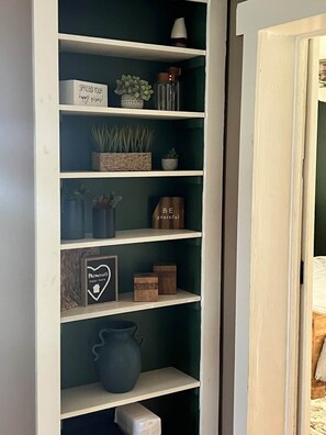 A white shelving unit with various decorative items including a sign that says 'SPREAD YOUR happy thoughts,' a small potted plant, a candle, and a framed picture with the text 'Memories made here.' - Quaint City Cottage just minutes from Killington (Rutland)