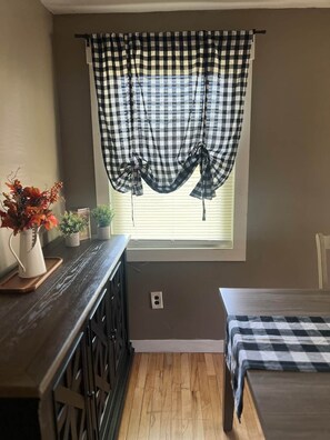 Une petite pièce avec une fenêtre ornée d’un rideau à carreaux, une commode en bois, une table de chevet et un lit recouvert d’une couverture à carreaux.