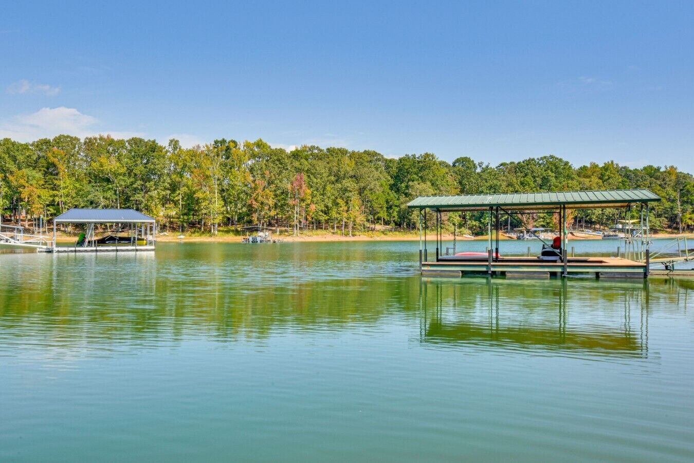 Un lac paisible où sont amarrés deux bateaux, avec un espace de pique-nique abrité et une rive bordée d’une forêt.