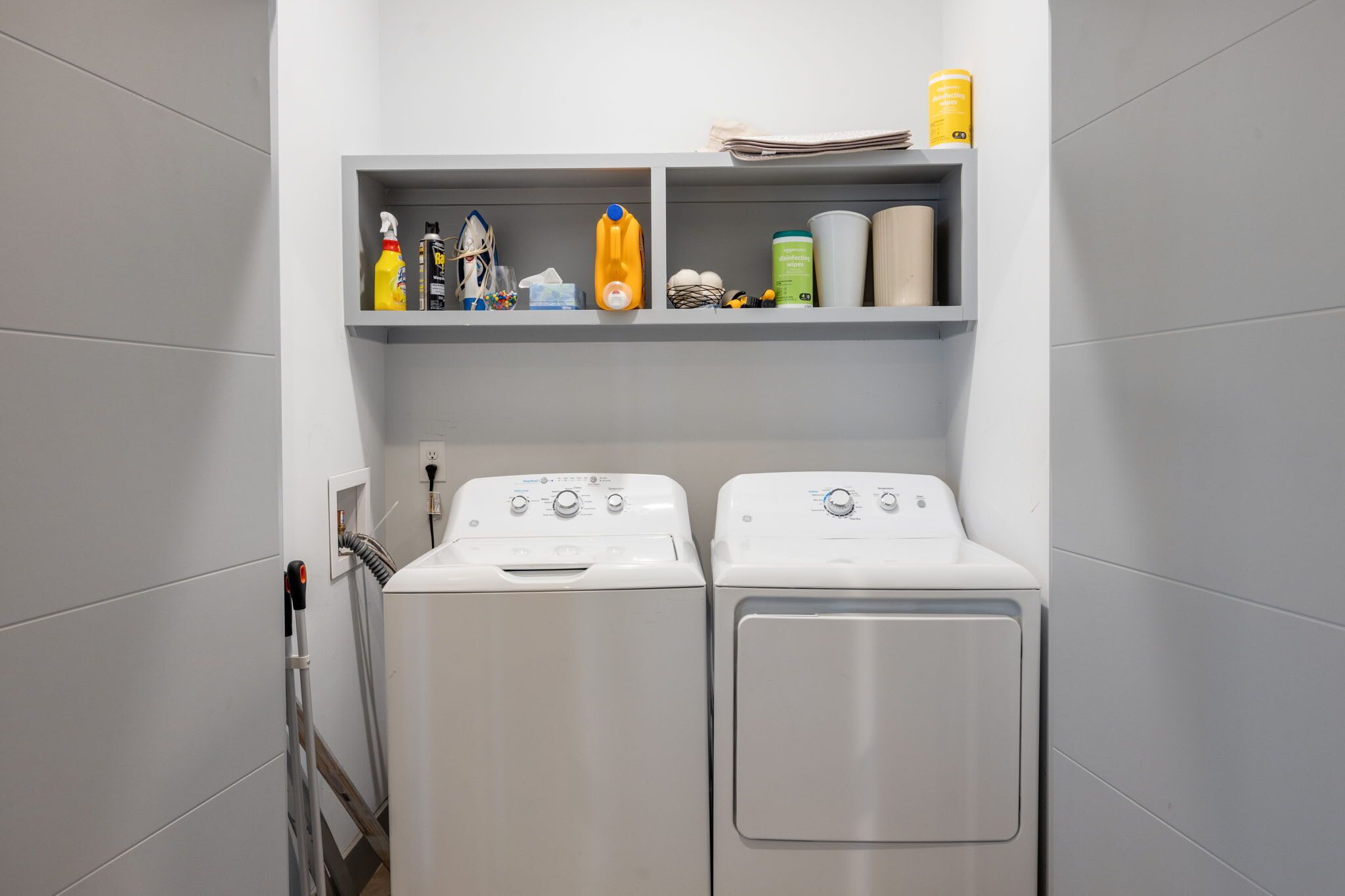 A laundry area with two front-loading washing machines and a shelf above containing cleaning supplies.