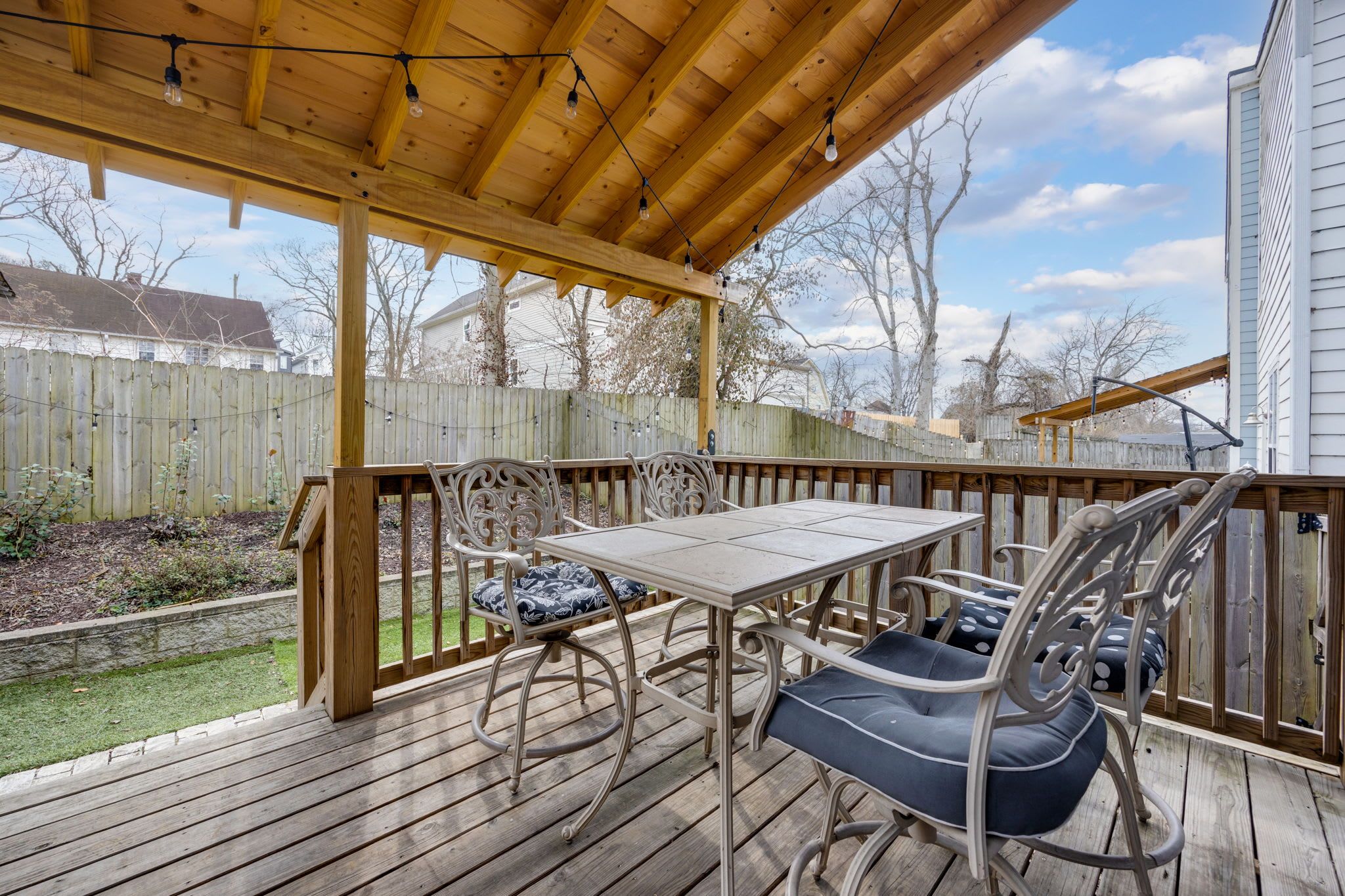 Une terrasse en bois avec une table et des chaises, une pergola illuminée de guirlandes lumineuses, et une vue sur le jardin et les maisons voisines.