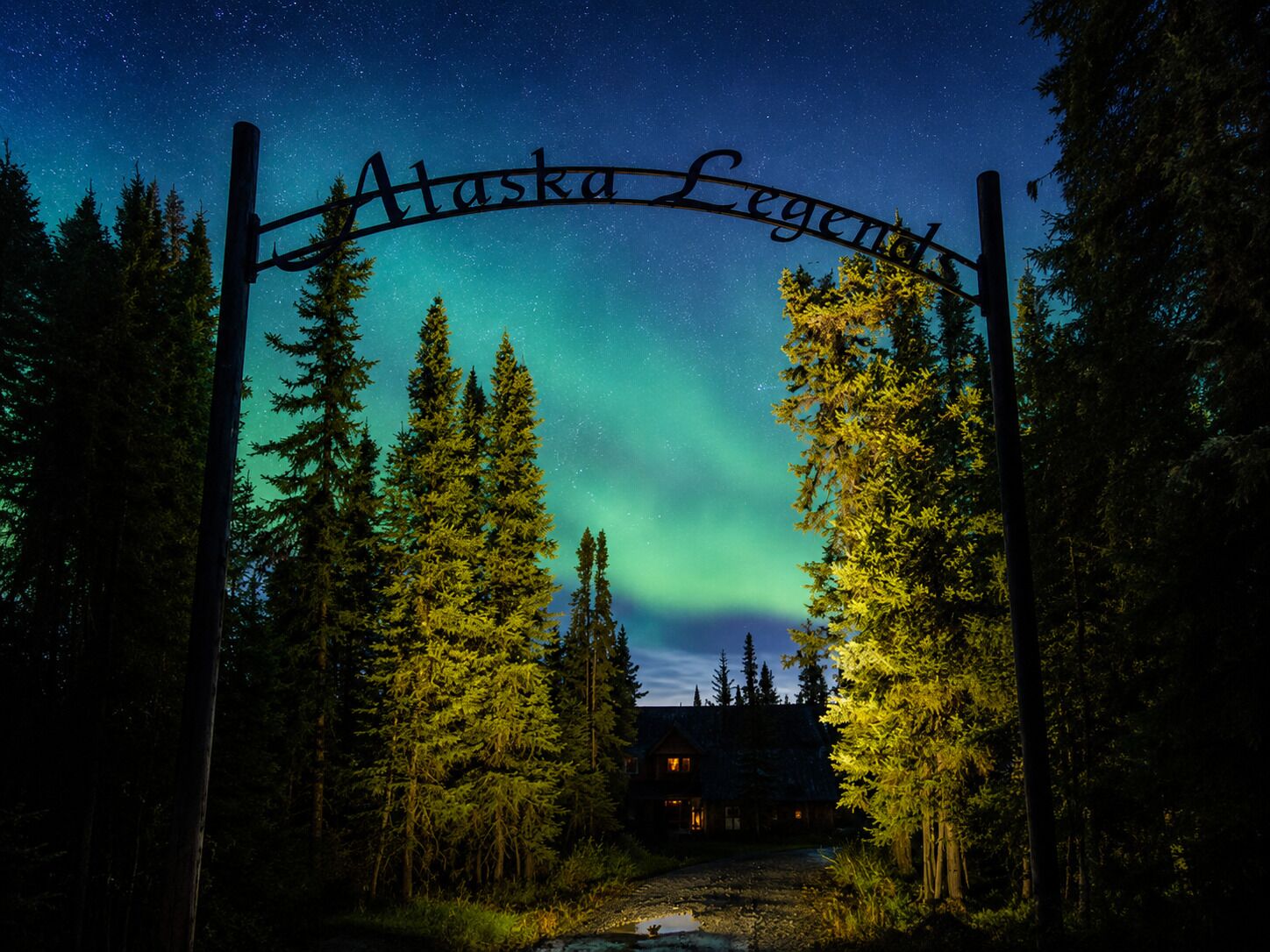 Un letrero de madera con la inscripción 'Alaska Legend', sobre un cielo nocturno con estrellas visibles y las auroras boreales.